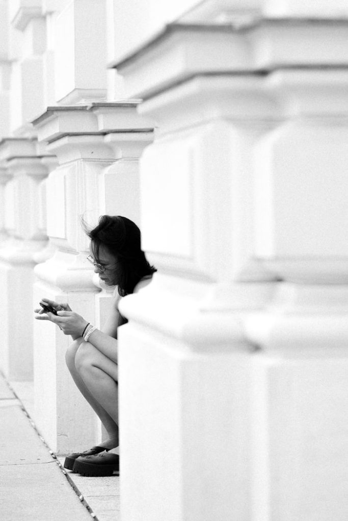 A woman crouched by a column in black and white, checking her phone.